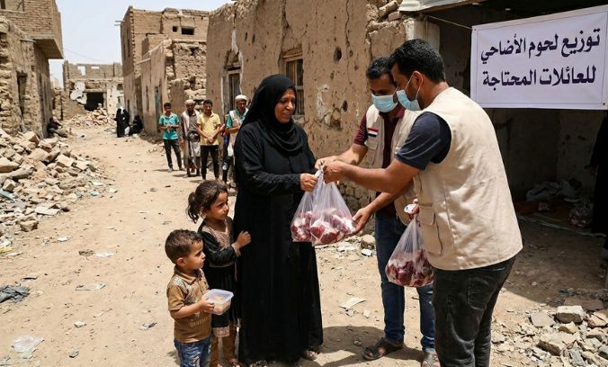 Two aid workers wearing face masks and vests distribute bags of Qurbani meat to a woman in a black abaya standing with two young children. They are on a dirt street in a conflict-affected area with damaged buildings and rubble. In the background, other people are waiting in line, and a banner with Arabic text is mounted on a nearby wall.