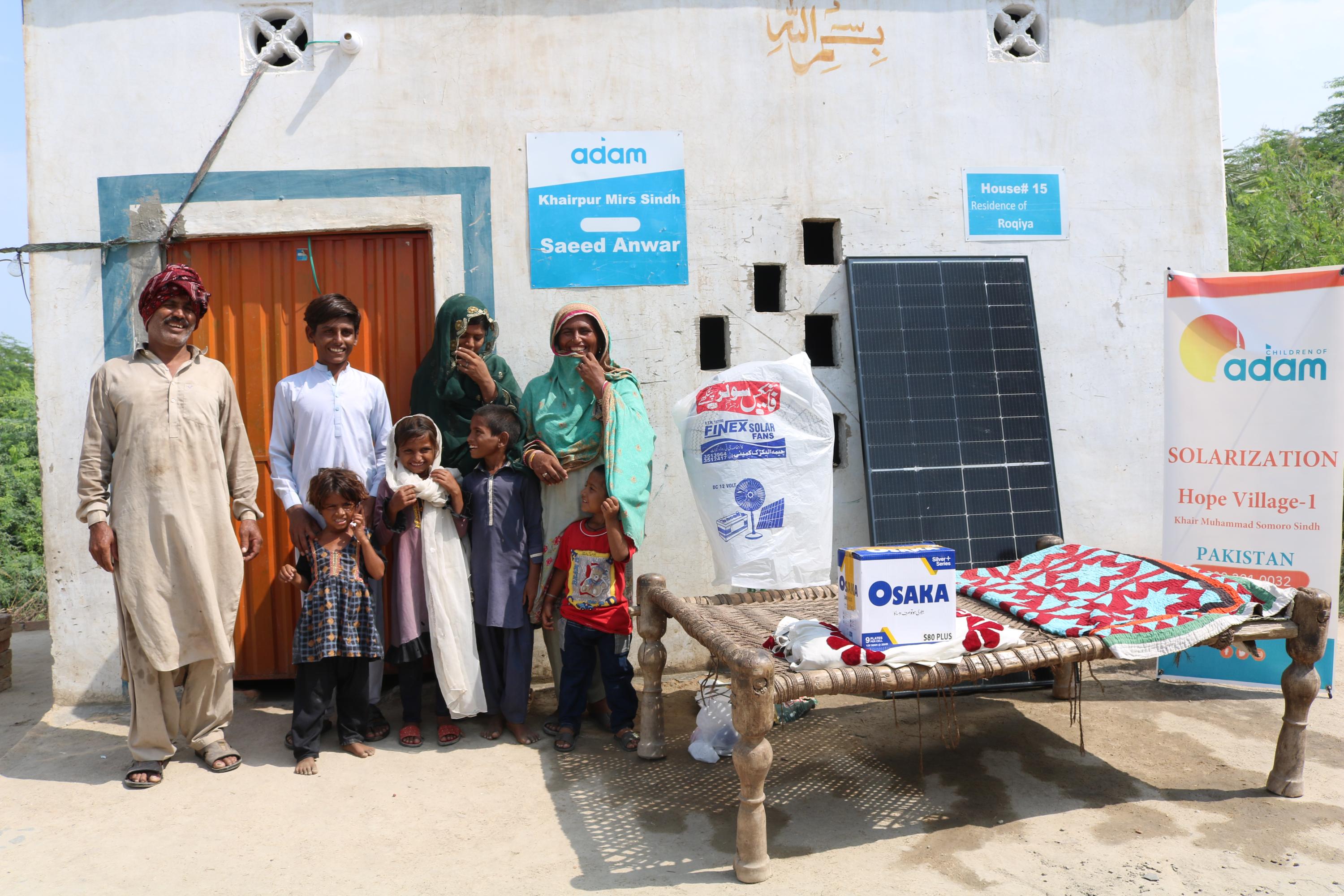 a smiling family poses outside their home in khairpur mirs, sindh, next to a solar panel, an osaka battery box, and a standing fan.