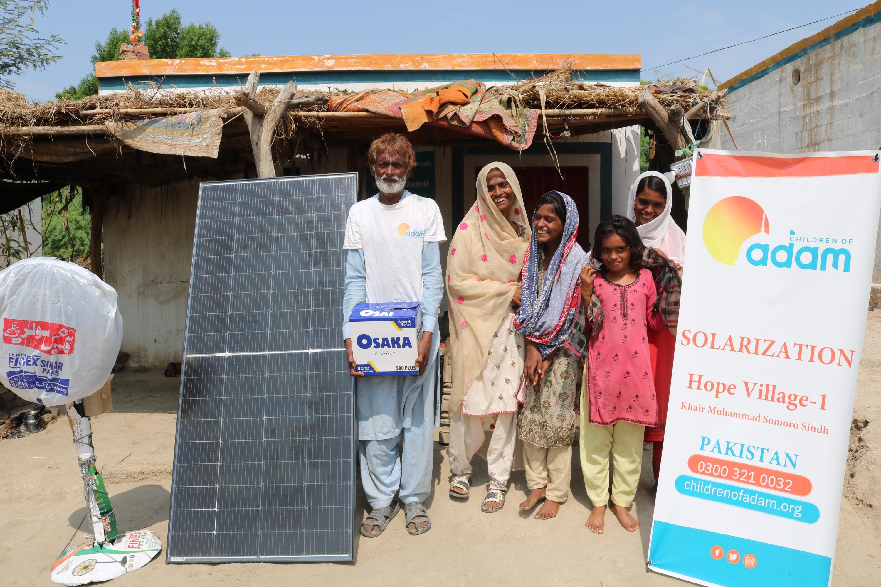 a smiling family poses outside their home in khairpur muhammad somoro, sindh, next to a solar panel, an osaka battery box, and a standing fan.