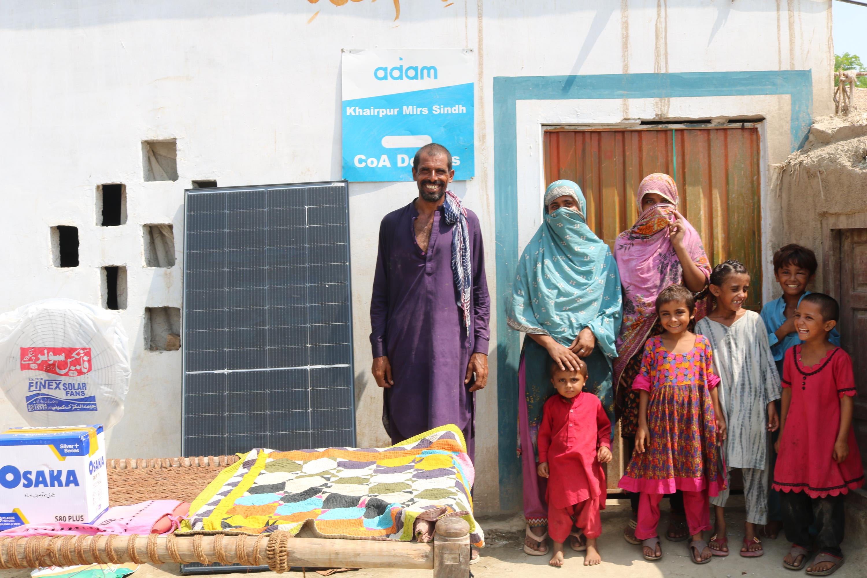 A smiling family poses outside their home in Khairpur Mirs, Sindh, next to a solar panel, an Osaka battery box, and a standing fan.