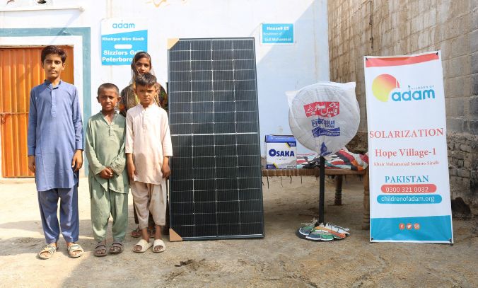 four children stand outside their home in khairpur mirs, sindh, next to a newly delivered solar panel, an osaka battery box, and a standing fan.