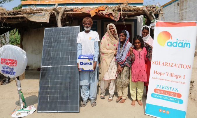 a smiling family poses outside their home in khairpur muhammad somoro, sindh, next to a solar panel, an osaka battery box, and a standing fan.