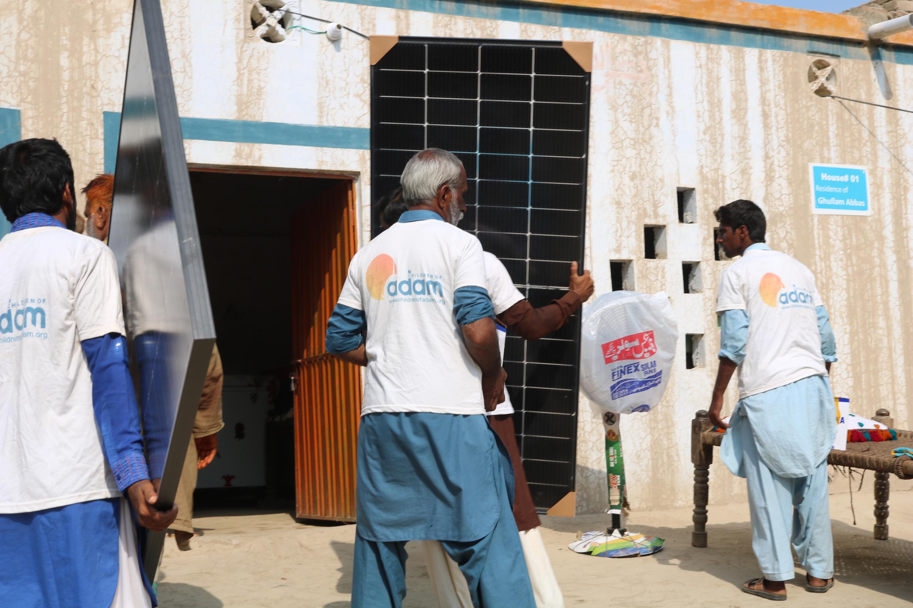 men wearing "children of adam" t-shirts carry and set up solar panels outside a house labeled "residence of ghullam abbas."